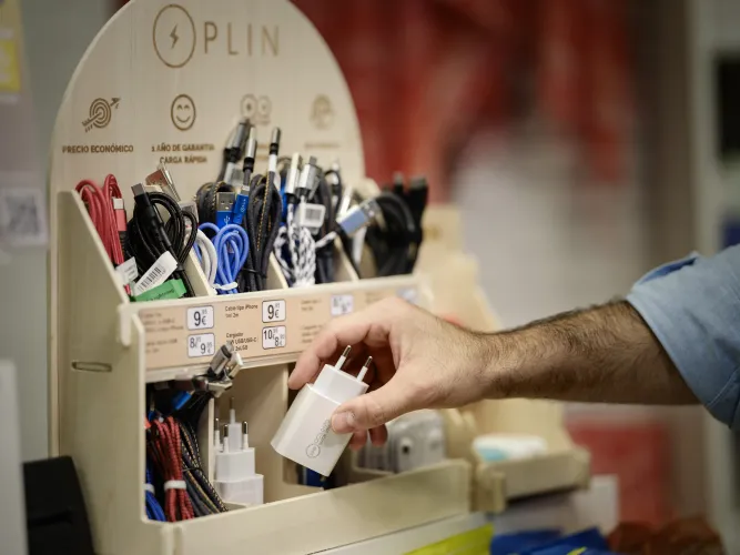 La mano de una persona coge un cargador de teléfono blanco de un expositor de cables de carga y dispositivos de colores en una tienda, con las etiquetas de precio a la vista.