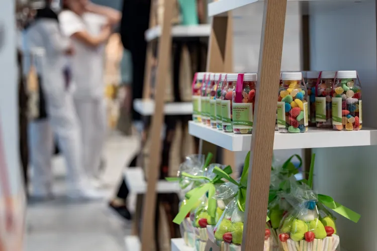 Estanterías llenas de tarros de caramelos de colores y bolsas atadas con cintas verdes. Al fondo se difuminan personas vestidas de blanco, creando un ambiente animado.