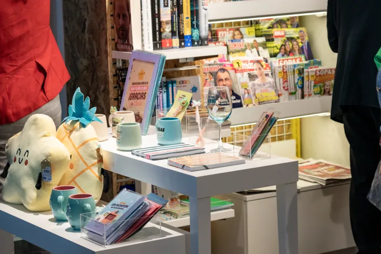 Una mesa expositora con peluches, tazas de colores, cuadernos y una copa de vino en una librería. Detrás, estanterías con revistas y libros. Luminoso y acogedor.