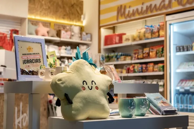 Un acogedor escaparate con un sonriente cojín en forma de estrella, rodeado de bonitas tazas y artículos de colores. Al fondo, estanterías con aperitivos y una nevera.