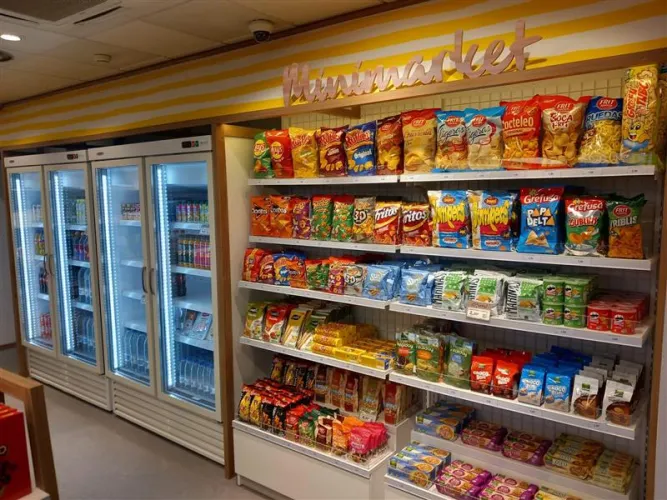 Un estante de minimercado con una variedad de papas fritas, snacks y barritas de cereales. Los refrigeradores a la izquierda contienen bebidas. Luminoso y organizado, evocando comodidad.