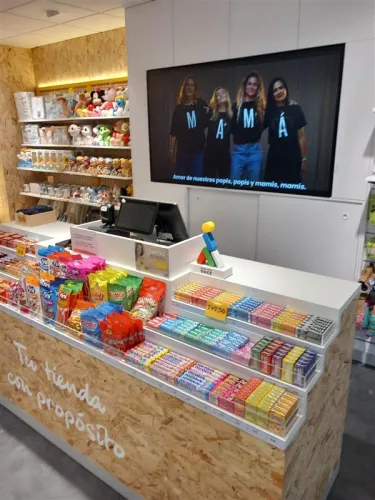 Interior de tienda moderno con una caja llena de dulces y coloridos snacks. Un letrero dice "Tu tienda con propósito". Los estantes exhiben peluches, y una pantalla superior muestra a cuatro personas con camisetas de "MAMÁ", lo que le da un toque cálido y familiar.