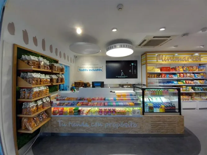 Minimercado luminoso con estanterías de pan, snacks y dulces. Un mostrador ofrece jugos frescos y ensaladas, creando un ambiente acogedor y vibrante.