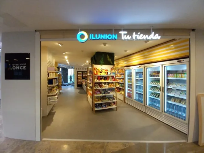 Entrada de tienda con cartel luminoso “ILUNION Tu tienda”, estanterías de juguetes y productos, neveras a la derecha, ambiente acogedor y organizado.