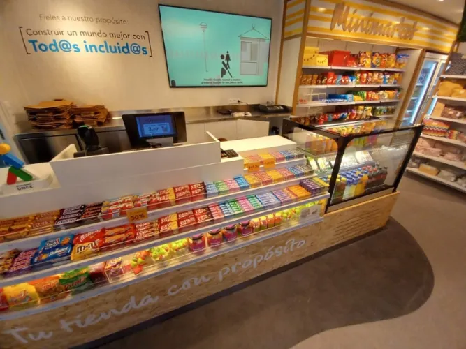 Dentro de un minimercado bien iluminado, con coloridos bocadillos y bebidas en un mostrador. Una pantalla digital muestra mensajes inclusivos. Ambiente acogedor y atractivo.