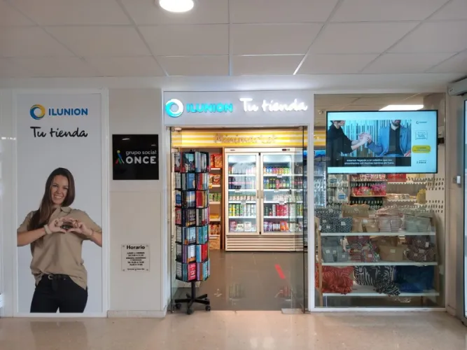 Entrada de una tienda ILUNION con letreros luminosos. Un expositor muestra diversos snacks y bebidas. Se reproduce un vídeo promocional en una pantalla.