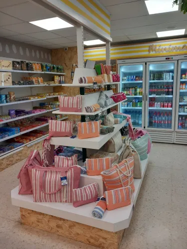 Exhibición de bolsas de rayas de colores y toallas enrolladas en un estante de una tienda de ILUNION. Detrás, estantes con refrigerios y bebidas refrigeradas.