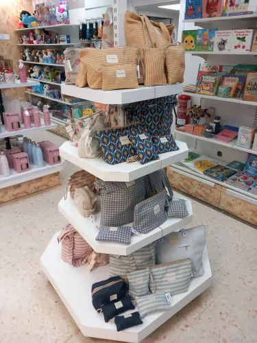 En una tienda, se exhiben diversos bolsos y carteras estampados en estantes hexagonales. Cerca, se ven libros y juguetes infantiles de colores vibrantes, creando un ambiente alegre y acogedor.