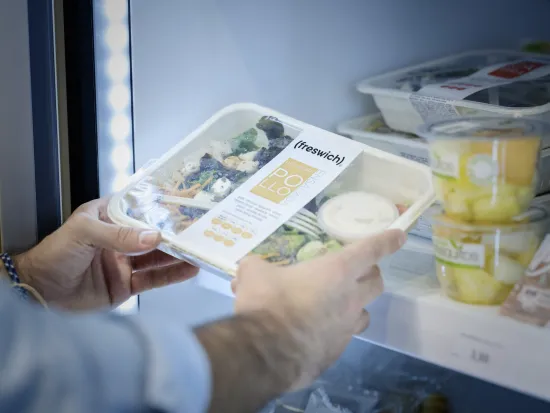 Unas manos sostienen una ensalada de aguacate y pollo envasada con la etiqueta «freswich» en un frigorífico. Cerca, hay vasos de fruta apilados. El entorno es luminoso y limpio.