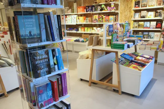 Una vibrante escena con estanterías de coloridos libros y juegos. En primer plano, una exposición de novelas; al fondo, libros infantiles y juguetes. Ambiente acogedor.