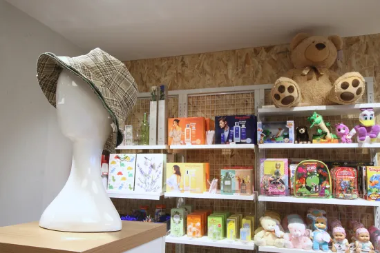 Una cabeza de maniquí con sombrero a cuadros se exhibe sobre un pedestal de madera. Detrás, unas estanterías repletas de juguetes de colores, juegos y grandes osos de peluche crean un ambiente lúdico.