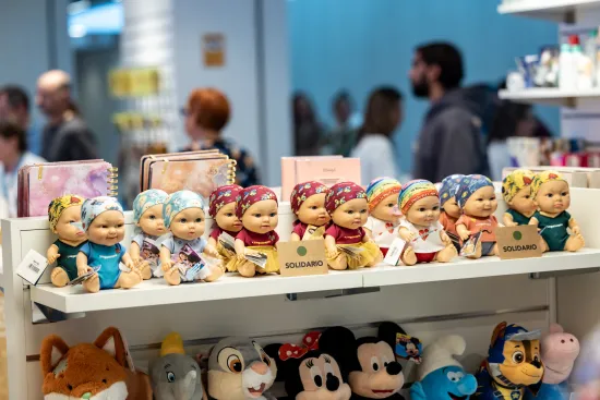 Una hilera de coloridas muñecas de bebé con diademas se exhibe en la estantería de una tienda. Debajo hay juguetes de peluche. En el fondo hay gente curioseando.