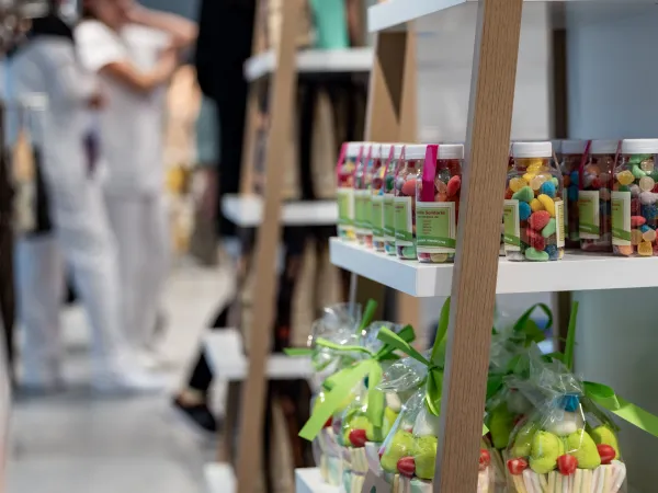 Estanterías llenas de tarros de caramelos de colores y bolsas atadas con cintas verdes. Al fondo se difuminan personas vestidas de blanco, creando un ambiente animado.