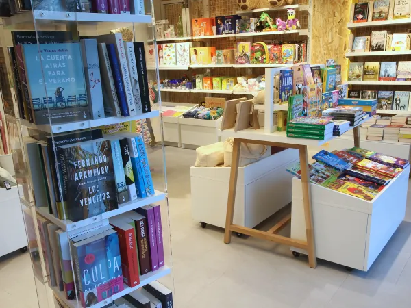 Una vibrante escena con estanterías de coloridos libros y juegos. En primer plano, una exposición de novelas; al fondo, libros infantiles y juguetes. Ambiente acogedor.