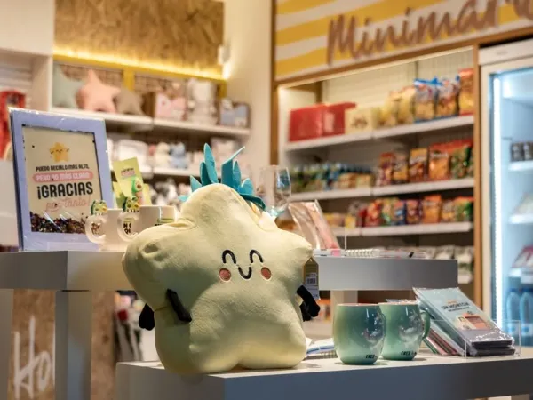 Un acogedor escaparate con un sonriente cojín en forma de estrella, rodeado de bonitas tazas y artículos de colores. Al fondo, estanterías con aperitivos y una nevera.