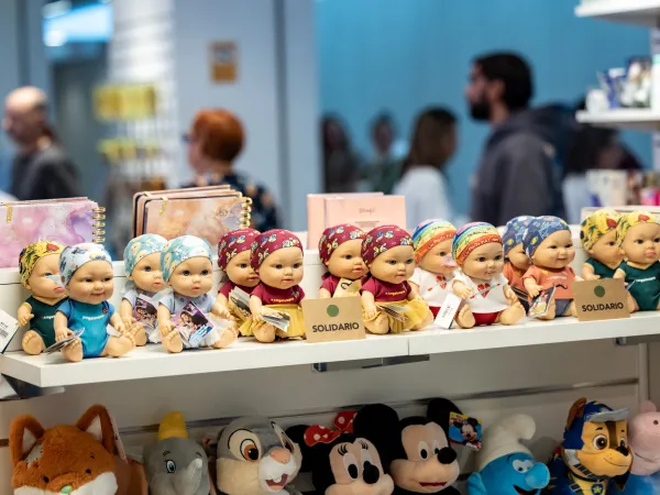 Una hilera de coloridas muñecas de bebé con diademas se exhibe en la estantería de una tienda. Debajo hay juguetes de peluche. En el fondo hay gente curioseando.