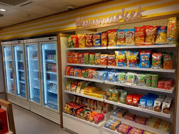 Un estante de minimercado con una variedad de papas fritas, snacks y barritas de cereales. Los refrigeradores a la izquierda contienen bebidas. Luminoso y organizado, evocando comodidad.