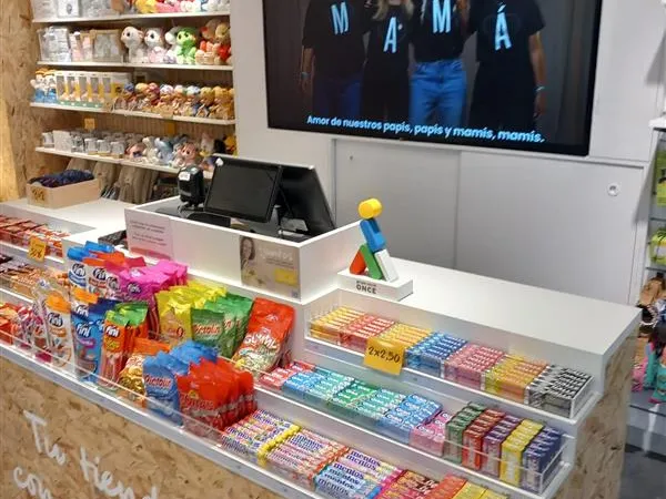Interior de tienda moderno con una caja llena de dulces y coloridos snacks. Un letrero dice "Tu tienda con propósito". Los estantes exhiben peluches, y una pantalla superior muestra a cuatro personas con camisetas de "MAMÁ", lo que le da un toque cálido y familiar.