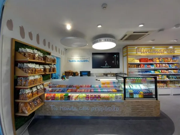 Minimercado luminoso con estanterías de pan, snacks y dulces. Un mostrador ofrece jugos frescos y ensaladas, creando un ambiente acogedor y vibrante.