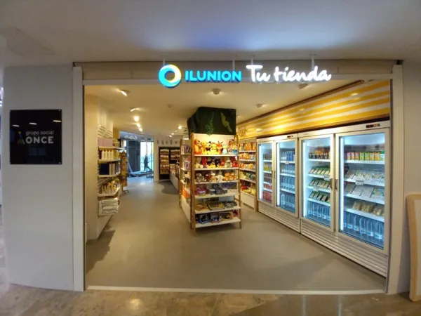 Entrada de tienda con cartel luminoso “ILUNION Tu tienda”, estanterías de juguetes y productos, neveras a la derecha, ambiente acogedor y organizado.