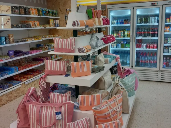 Exhibición de bolsas de rayas de colores y toallas enrolladas en un estante de una tienda de ILUNION. Detrás, estantes con refrigerios y bebidas refrigeradas.