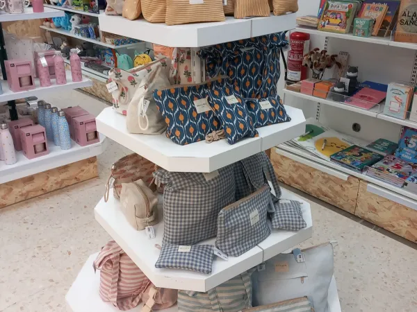 En una tienda, se exhiben diversos bolsos y carteras estampados en estantes hexagonales. Cerca, se ven libros y juguetes infantiles de colores vibrantes, creando un ambiente alegre y acogedor.
